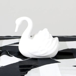 Könnte beinhalten: Eine weiße Keramik-Schwanfigur mit gebogenem Hals und detaillierten Federn. Der Schwan steht vor einem schwarz-weiß gemusterten Hintergrund. Ein dekoratives Objekt für die Inneneinrichtung, etwa 15 cm lang.