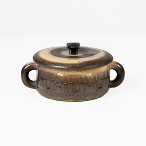 Può includere: Un piatto coperto in ceramica marrone e beige con un pomo nero sul coperchio. Il piatto ha un motivo intrecciato e un bordo verde.