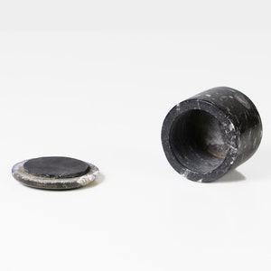 May include: A black marble container with a matching lid. The cylindrical container is open, revealing a hollow interior. The lid is a flat, circular piece. The marble has a speckled pattern.