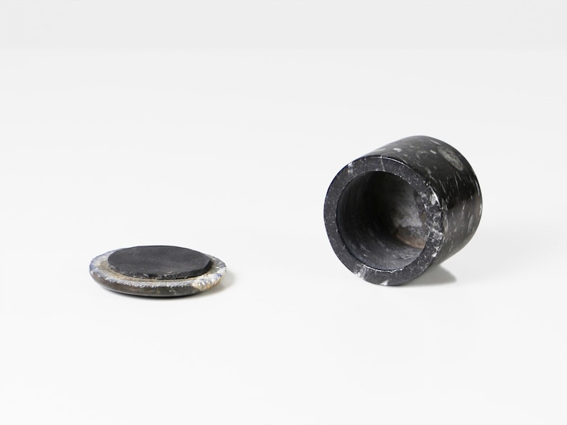 May include: A black marble container with a matching lid. The cylindrical container is open, revealing a hollow interior. The lid is a flat, circular piece. The marble has a speckled pattern.