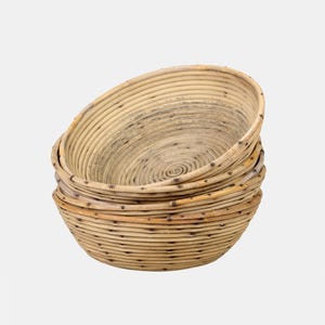 Op de afbeelding: Een stapel van vier ronde, lichtbruine rotan manden. De manden zijn geweven in een spiraalpatroon, met een donkere vlek in het midden van elk. De zijkanten van de manden zijn gemaakt van horizontale rotan strips.