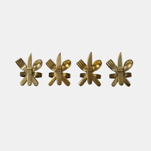 Può includere: Quattro anelli portatovaglioli in metallo color oro. Ogni anello presenta una forchetta, un coltello e un cucchiaio disposti in un motivo decorativo. Le posate sono collegate da un anello circolare, creando un accento unico per la tavola.