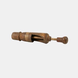 Op de afbeelding: Een vintage houten notenkraker met een cilindervormig lichaam in een verweerde bruine afwerking, een centrale open gleuf en een slank plunjermechanisme met een ronde knopgreep die aan één uiteinde van het gereedschap uitsteekt voor bediening.