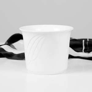 Peut inclure: Pot de fleurs en céramique blanche avec un motif incurvé sur le côté. Le pot a un bord évasé et repose sur une surface blanche. Un tissu à rayures noires et blanches est en arrière-plan.