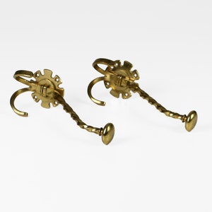 Op de afbeelding: Een paar gouden metalen kapstokhaken met een gedraaid ontwerp. Elke haak heeft een decoratieve ronde basis met een reliëf.