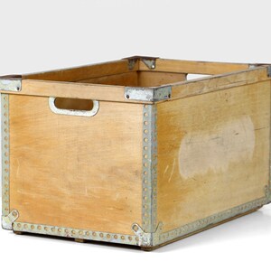 May include: A wooden storage crate with metal accents. The crate is made of light wood and has a rectangular shape. The metal accents are silver and are located around the edges of the crate.
