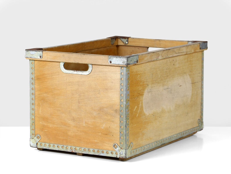 May include: A wooden storage crate with metal accents. The crate is made of light wood and has a rectangular shape. The metal accents are silver and are located around the edges of the crate.