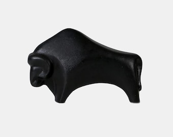 May include: A solid black sculpture of a bull in a minimalist style. The bull is depicted in a standing position, with a curved back and a defined head. The sculpture has a smooth, matte finish and is likely made of a solid material.