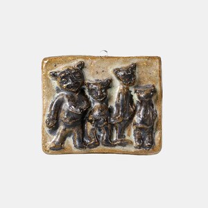 May include: A ceramic wall hanging featuring a relief of four stylized bear-like figures. The figures are dark brown with lighter accents, set against a tan background. The piece has a square shape and a loop for hanging.