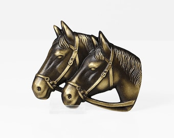 May include: Two gold-toned horse head sculptures with bridles. The horses are facing the same direction, with the horse in the back slightly overlapping the horse in the front.