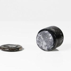 May include: A black marble tumbler and coaster set. The tumbler is cylindrical, with a speckled pattern. The coaster is round and flat, with a similar speckled pattern. Both pieces are made of a dark, natural stone.