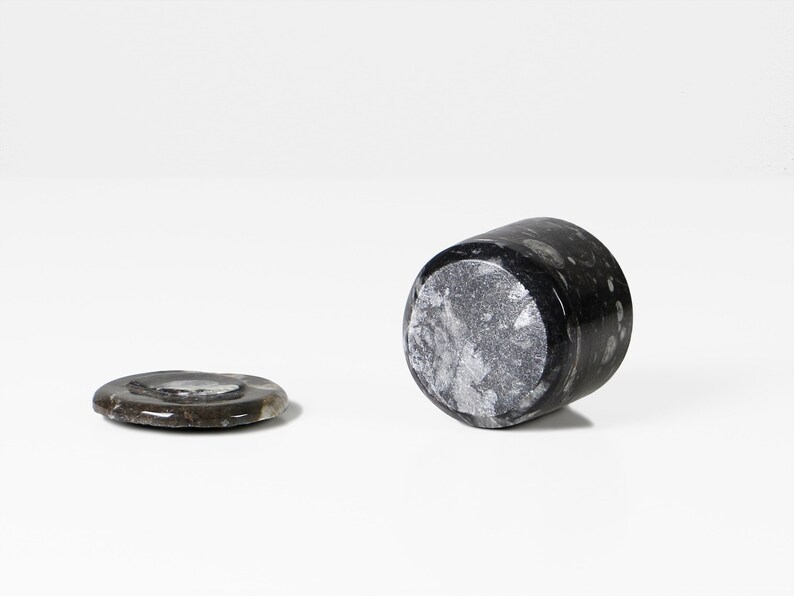 May include: A black marble tumbler and coaster set. The tumbler is cylindrical, with a speckled pattern. The coaster is round and flat, with a similar speckled pattern. Both pieces are made of a dark, natural stone.