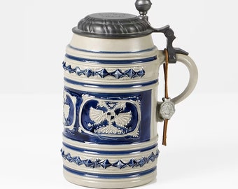 May include: A ceramic beer stein with a blue and white design. The stein has a pewter lid with a knob on top and a leather strap with a pewter charm. The stein is decorated with a coat of arms featuring a double-headed eagle.