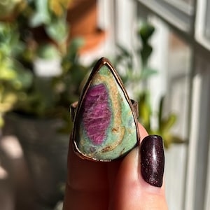 May include: A copper ring with a teardrop-shaped green and pink gemstone. The gemstone has a swirling pattern of green, pink, and gold.