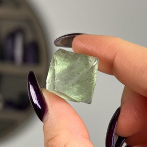 May include: A light green fluorite crystal with a cube shape. The crystal has a smooth, polished surface and is translucent.