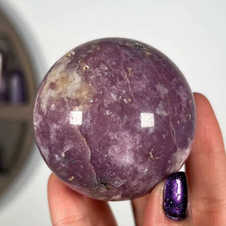 May include: A large, polished, purple lepidolite sphere with a smooth, shiny surface. The sphere has a light purple color with white and gray inclusions.