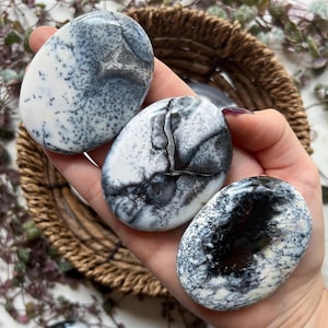 May include: Three polished dendritic opal stones with a mix of white, grey, and black patterns. The stones are oval-shaped and held in a hand, with a woven basket in the background. The stones have a smooth, glossy finish.