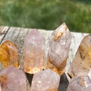 May include: A collection of seven clear quartz crystal points with inclusions of golden rutile and pink hues. The points are arranged on a wooden surface.