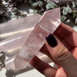 Può includere: Una bacchetta di cristallo di quarzo rosa trasparente e rosa con una punta. Il cristallo è tenuto in una mano, con altri cristalli simili visibili sullo sfondo. Il cristallo ha un aspetto traslucido ed è di colore rosa chiaro.