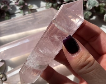 May include: A clear, pink rose quartz crystal wand with a pointed tip. The crystal is held in a hand, with other similar crystals visible in the background. The crystal has a translucent appearance and is a light pink color.