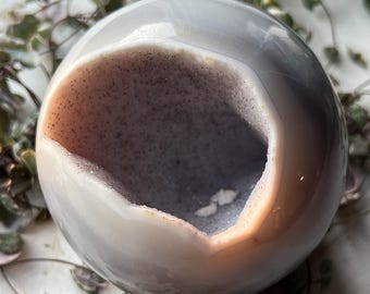 May include: A white agate geode with a large opening revealing a purple and white interior. The geode is smooth and polished, with a natural, organic shape.