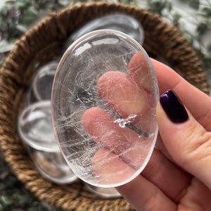 May include: A clear quartz crystal palm stone with a smooth, polished surface. The stone has a natural, cloudy appearance with visible inclusions.