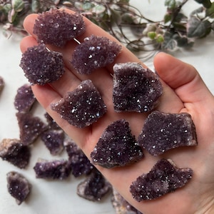 Op de afbeelding: Een verzameling donkerpaarse amethistkristalclusters. De kristallen hebben een glinsterend, getextureerd oppervlak en worden gepresenteerd op een lichte achtergrond. De amethiststukken variëren in grootte en vorm en tonen natuurlijke formaties.