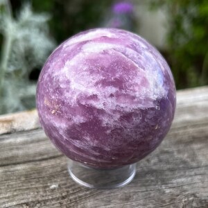May include: A large, polished amethyst sphere with a swirling purple and white pattern. The sphere is displayed on a clear stand.