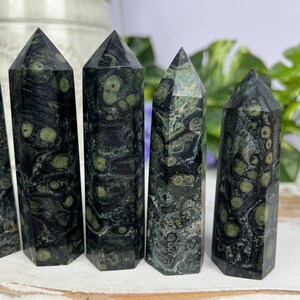Pode incluir: Cinco pontas de cristal de pedra verde e preta salpicada, tamb&eacute;m conhecidas como aventurina verde, dispostas em fila sobre uma superf&iacute;cie de madeira branca.
