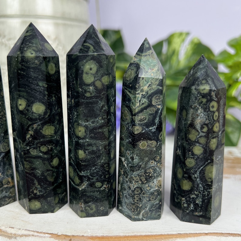 Pode incluir: Cinco pontas de cristal de pedra verde e preta salpicada, tamb&eacute;m conhecidas como aventurina verde, dispostas em fila sobre uma superf&iacute;cie de madeira branca.