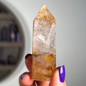 May include: A clear quartz crystal point with golden inclusions. The crystal is approximately 2 inches tall and has a pointed tip. The crystal is held in a hand for scale.