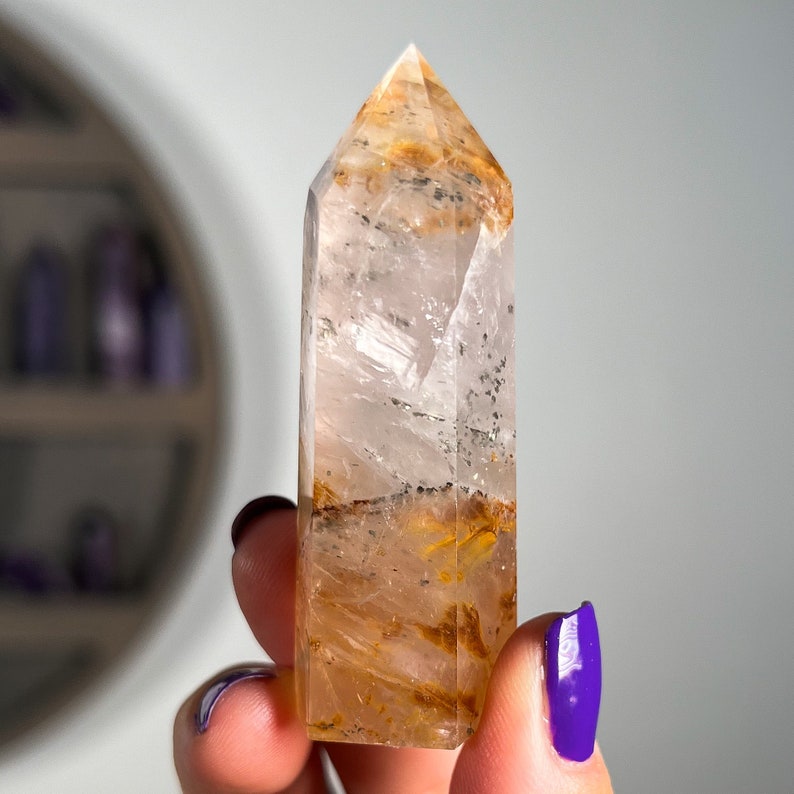 May include: A clear quartz crystal point with golden inclusions. The crystal is approximately 2 inches tall and has a pointed tip. The crystal is held in a hand for scale.