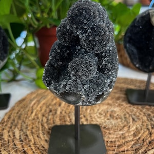May include: A dark black druzy mineral specimen featuring multiple rounded, crystalline clusters with a sparkling, textured surface. The mineral is mounted on a thin, vertical metal rod attached to a square, flat black metal base for display.
