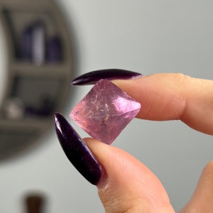 May include: A pink fluorite crystal with a natural, faceted shape. The crystal is clear and has a slight purple hue.