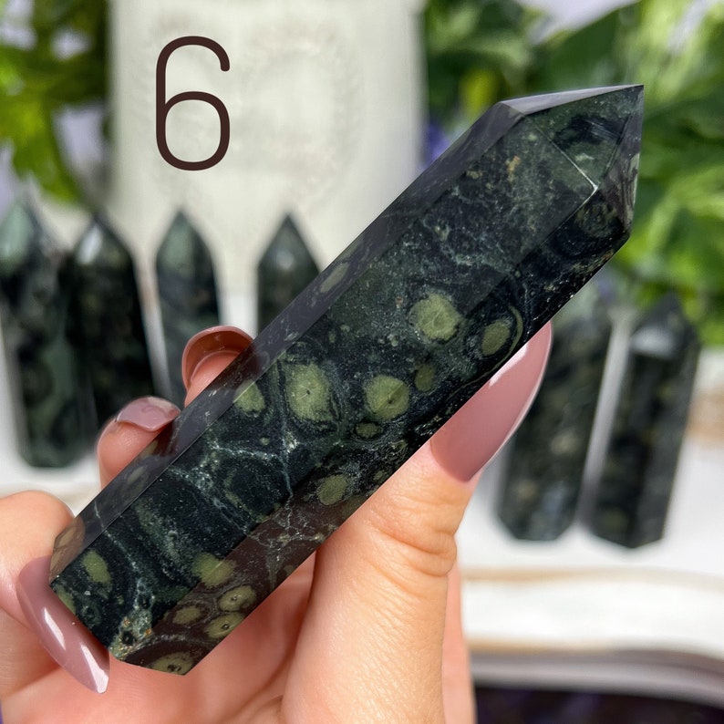 Pode incluir: Uma ponta de pedra verde e preta salpicada, tamb&eacute;m conhecida como varinha de cristal, &eacute; segurada na m&atilde;o. A pedra &eacute; polida e tem uma superf&iacute;cie lisa. A ponta tem aproximadamente 15 cm de comprimento.