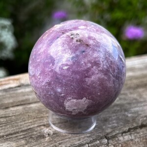 Puede incluir: Una gema esf&eacute;rica pulida, en tonos lavanda y p&uacute;rpura, sobre un soporte de acr&iacute;lico transparente. El orbe tiene una superficie texturizada con sutiles variaciones de color. El fondo presenta vegetaci&oacute;n borrosa.