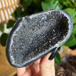 May include: A polished, oval-shaped geode with a dark gray exterior and a sparkling, crystal-filled interior. The geode's outer edge has a subtle blue-gray hue, and the inner cavity is filled with tiny, glittering crystals.