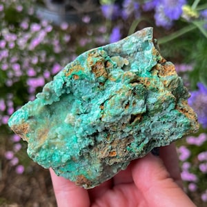 May include: A green and brown rough-cut crystal with a textured surface. The crystal has a variety of shades of green and brown, and appears to be a natural formation.