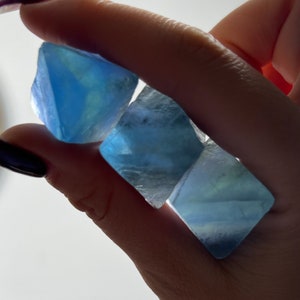 Puede incluir: Tres cristales de fluorita azules, cada uno con un tono de azul diferente, se sostienen en una mano. Los cristales son lisos y tienen un ligero brillo.