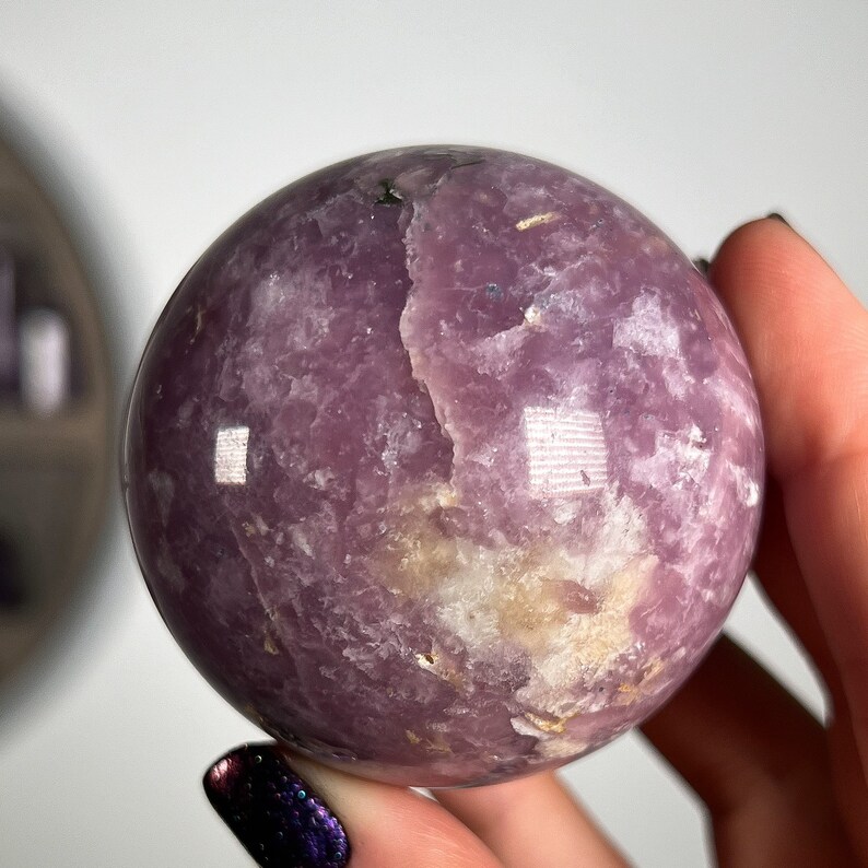 May include: A polished purple and white Lepidolite sphere crystal ball. The sphere has a smooth, polished surface and a natural, cloudy appearance.