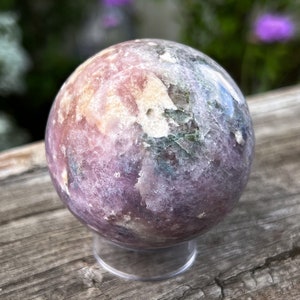 Puede incluir: Una gema esf&eacute;rica pulida, con una mezcla de colores lavanda, rosa y verde. La esfera se apoya en un soporte transparente y circular, resaltando sus texturas naturales. El fondo borroso destaca la esfera de cristal, un objeto decorativo.