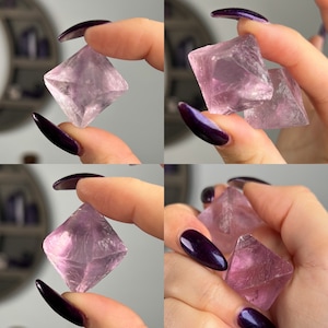 May include: Two pink fluorite crystals, one larger than the other, are held in a hand. The crystals are clear and have a slight purple hue. They are both octahedral in shape.