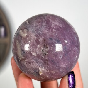 Puede incluir: Una gema esf&eacute;rica pulida, en tonos lavanda y p&uacute;rpura. El orbe tiene una superficie lisa y reflectante con variaciones naturales de color y textura. La gema se sostiene en una mano sobre un fondo blanco.
