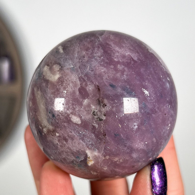 Puede incluir: Una gema esf&eacute;rica pulida, en tonos lavanda y p&uacute;rpura. El orbe tiene una superficie lisa y reflectante con variaciones naturales de color y textura. La gema se sostiene en una mano sobre un fondo blanco.