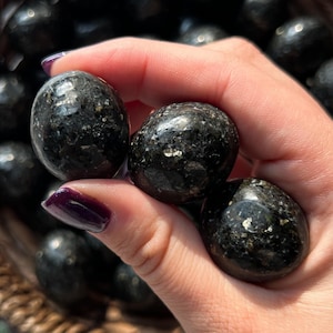 May include: A hand holds several polished black stones with flecks of gold. The stones are smooth and rounded, with a glossy finish. The background is blurred, suggesting a collection of similar stones.