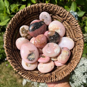 Peut inclure: Un panier en osier rempli de pierres d'opale rose polies. Les pierres, de tailles variées, présentent des marques roses, marron et noires. Le panier est en matériau tissé brun clair, avec un fond de feuillage vert flou.