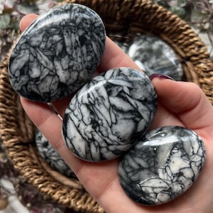 Peut inclure: Quatre pierres polies de forme ovale, présentant un motif noir et blanc saisissant, ressemblant à un réseau de veines ou de fissures. Les pierres sont tenues dans un panier tressé, mettant en valeur leur beauté naturelle et leurs textures uniques. Les pierres sont lisses et brillantes.