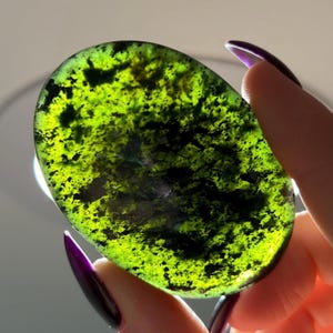 May include: An oval-shaped polished gemstone featuring a vibrant translucent lime green base color with deep forest green and black mottled inclusions throughout the surface, illuminated from behind to highlight the internal color variations.