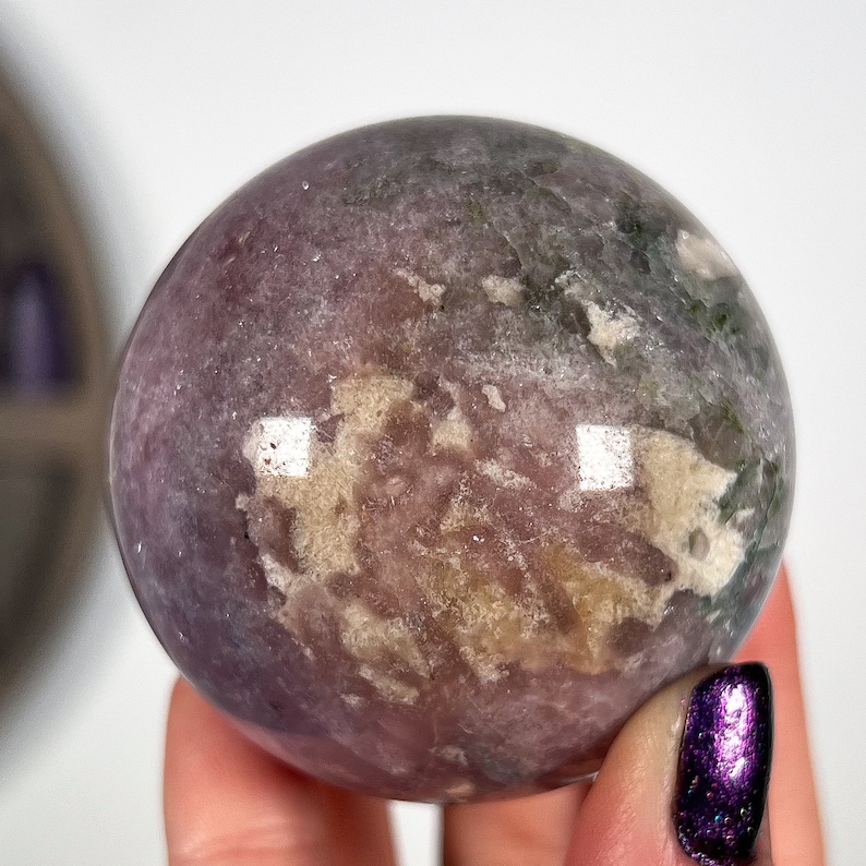 Puede incluir: Una gema esf&eacute;rica pulida, con tonos morados, verdes y crema. La esfera tiene una superficie lisa y reflectante con variaciones naturales de color y textura. La gema se sostiene en una mano, sobre un fondo blanco.