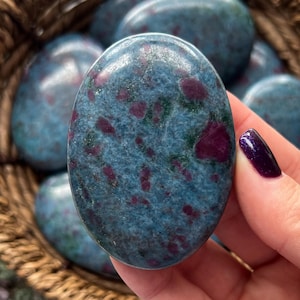 May include: A smooth, oval-shaped blue and red gemstone with a speckled pattern. The stone is polished and has a glossy finish.